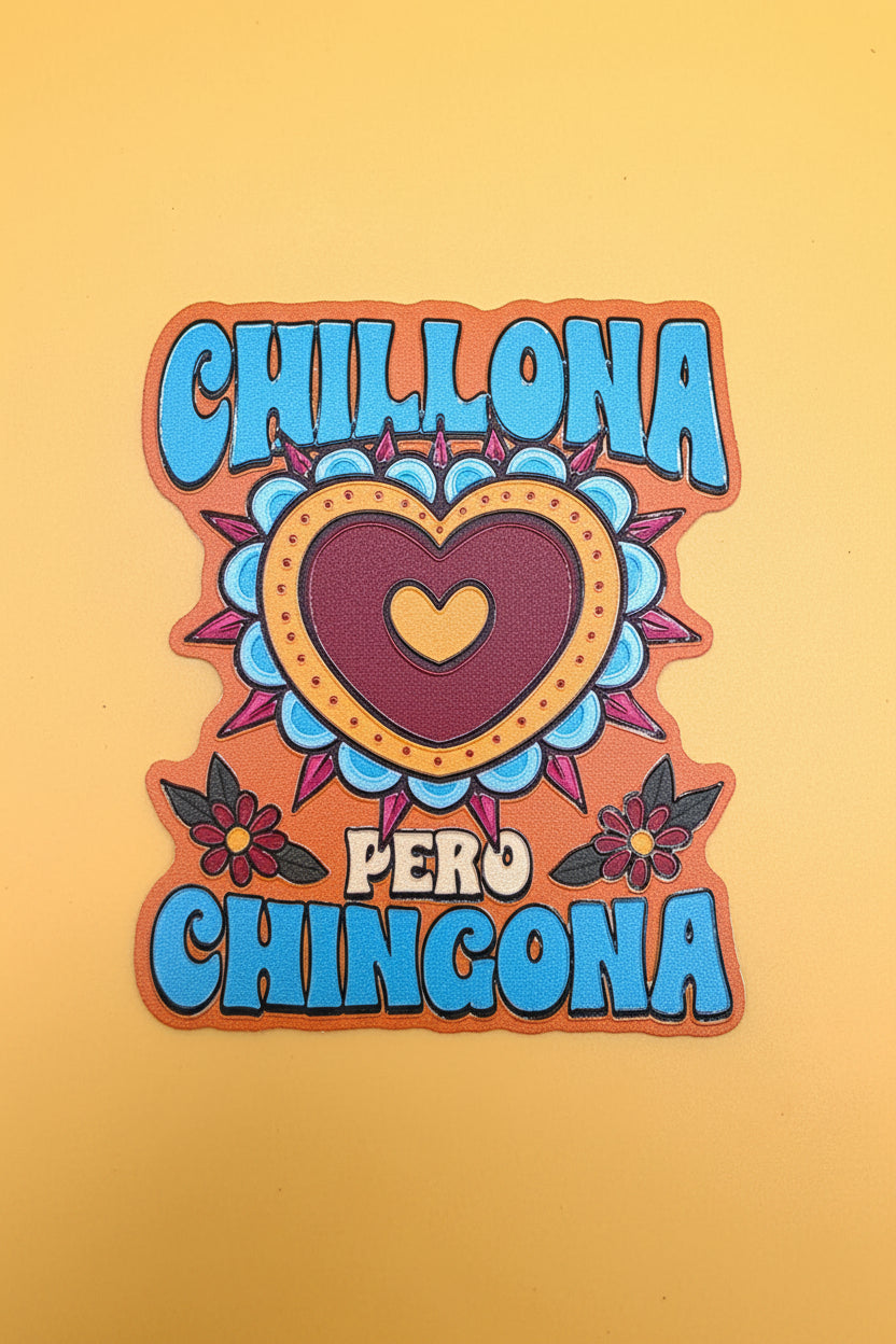 A die-cut decorative sticker or patch with the words “CHILLONA PERO CHINGONA” in large blue letters on an orange border, surrounding a central heart design in maroon, yellow, blue, and pink on a yellow background.
