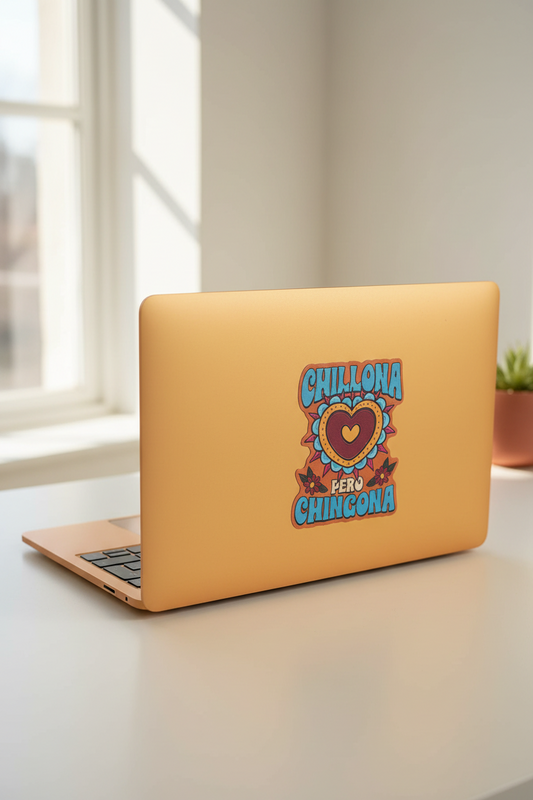 A closed laptop in a warm peachy-orange finish sits on a light tabletop near a window. The back of the laptop has a colorful graphic sticker with the words “CHILLONA BERO CHINGONA” around a heart design.