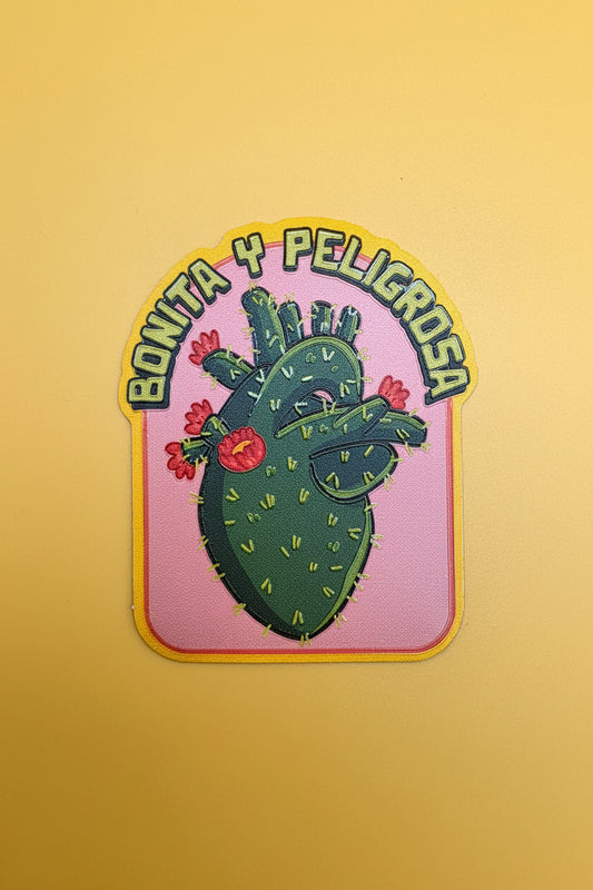 A die-cut sticker featuring a stylized green cactus with red flowers on a pink background, with the Spanish text 'BONITA Y PELIGROSA' in green outlined lettering around the top. The sticker is photographed against a solid pale yellow background.